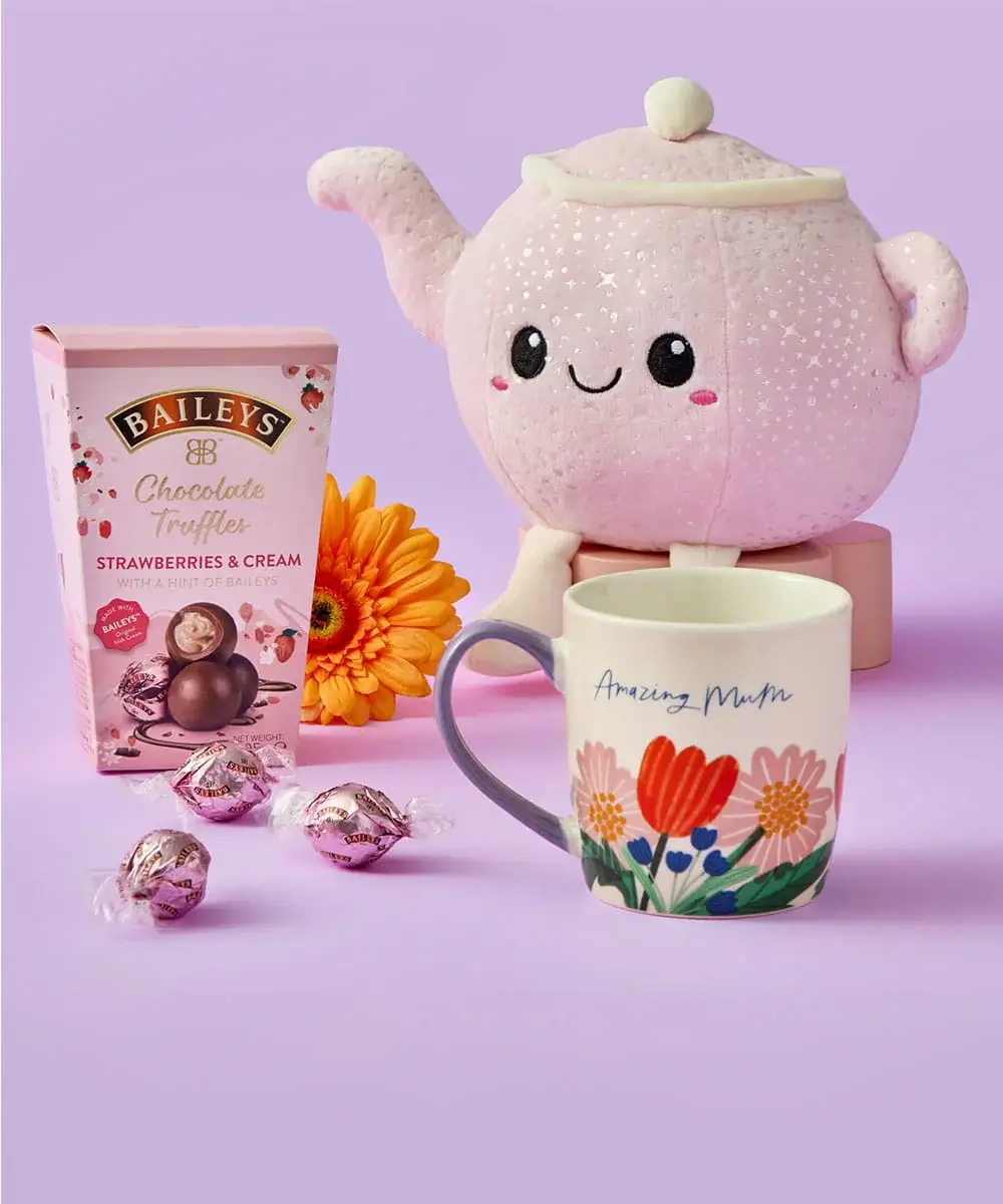 A Mother’s Day gift set featuring a pink teapot plush toy, a floral Amazing Mum mug, Baileys strawberries and cream truffles, and a bright orange flower arranged on a pastel purple background.